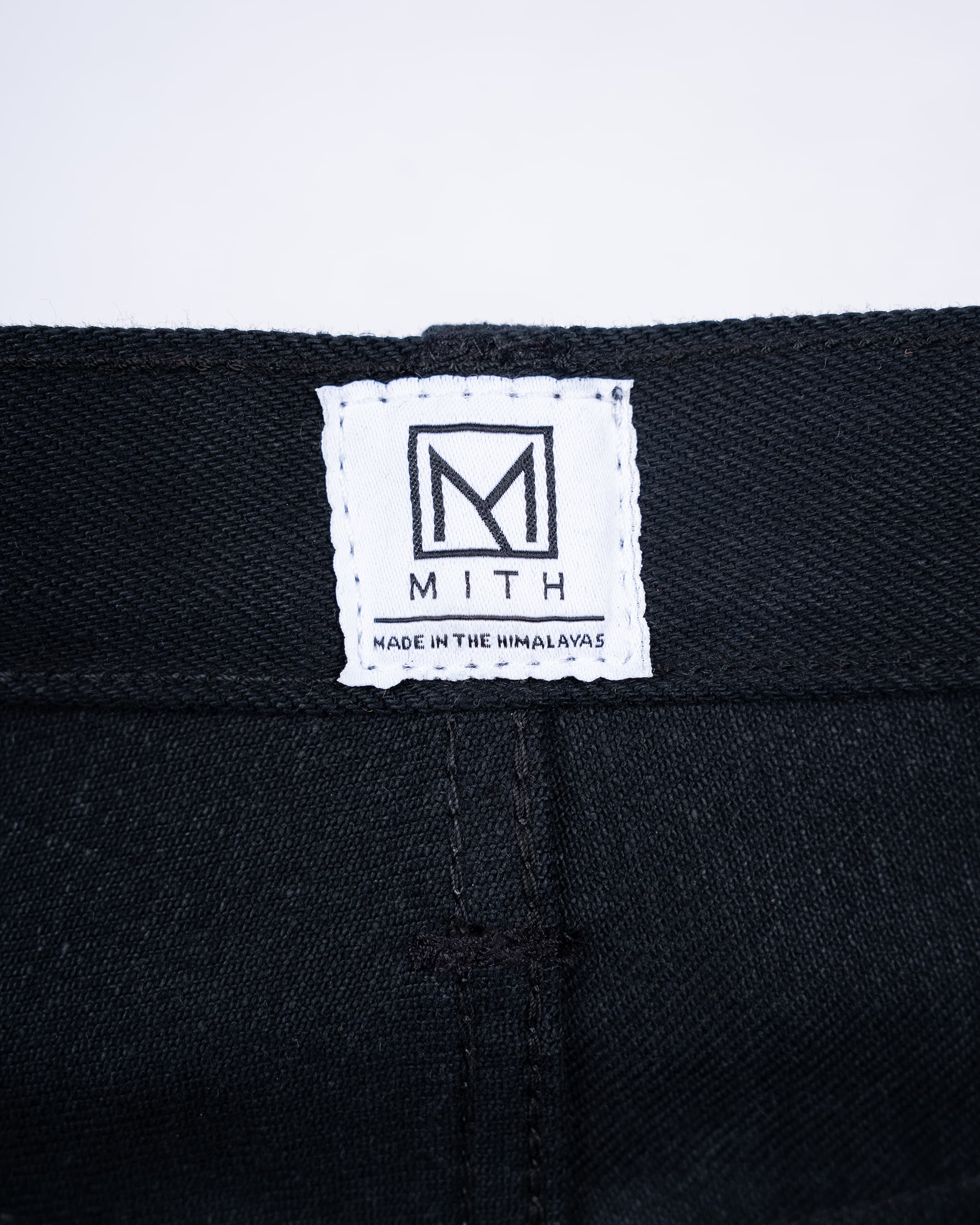 Close-up of a 17oz black selvedge denim waistband with a white sewn-in label featuring the MITH logo and 'MADE IN THE HIMALAYAS'.